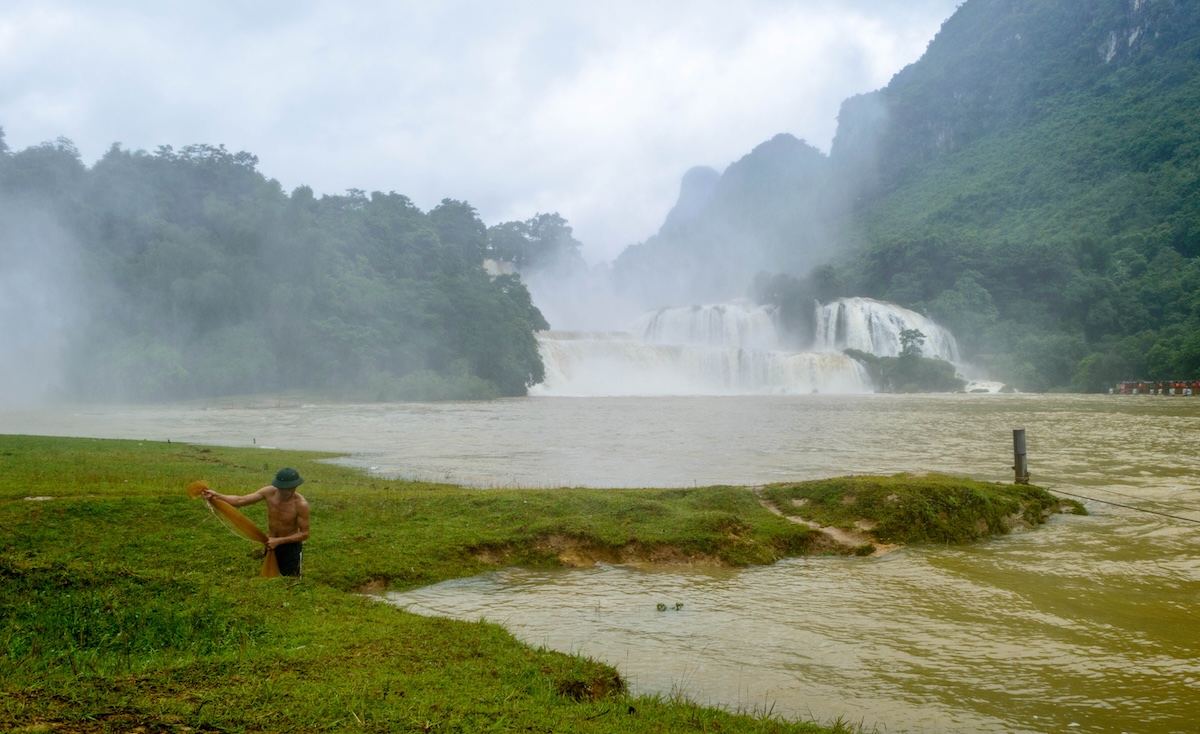 Een visser met een net op de grasoever van de rivier, met de Ban Gioc Waterval in de nevel op de achtergrond