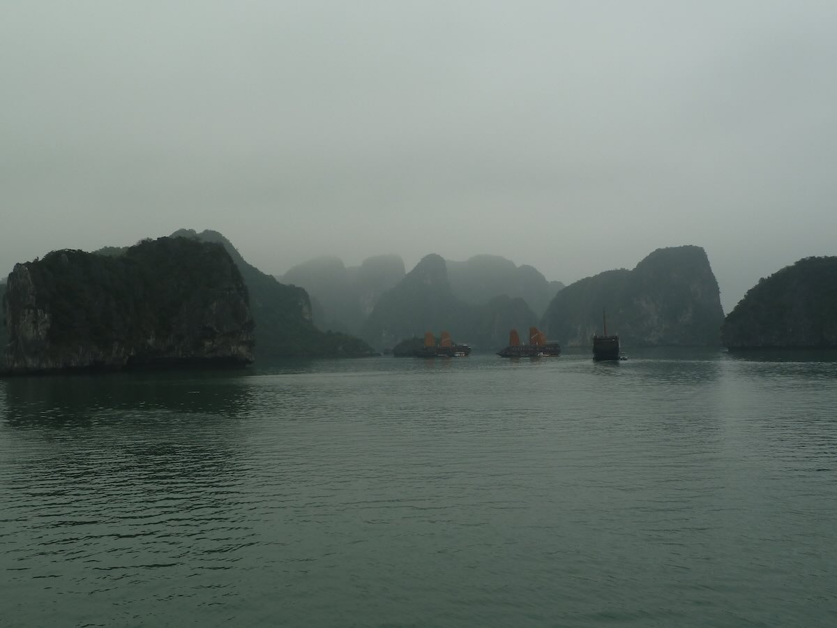 Jonken met oranje zeilen varen in de mist tussen de kalksteenrotsen van Ha Long Bay