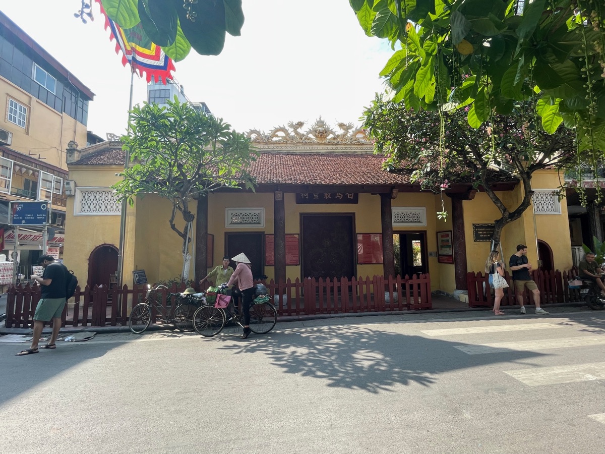 De gele gevel van de Bach Ma-tempel in Hàng Buồm, met een fietser en voetgangers ervoor
