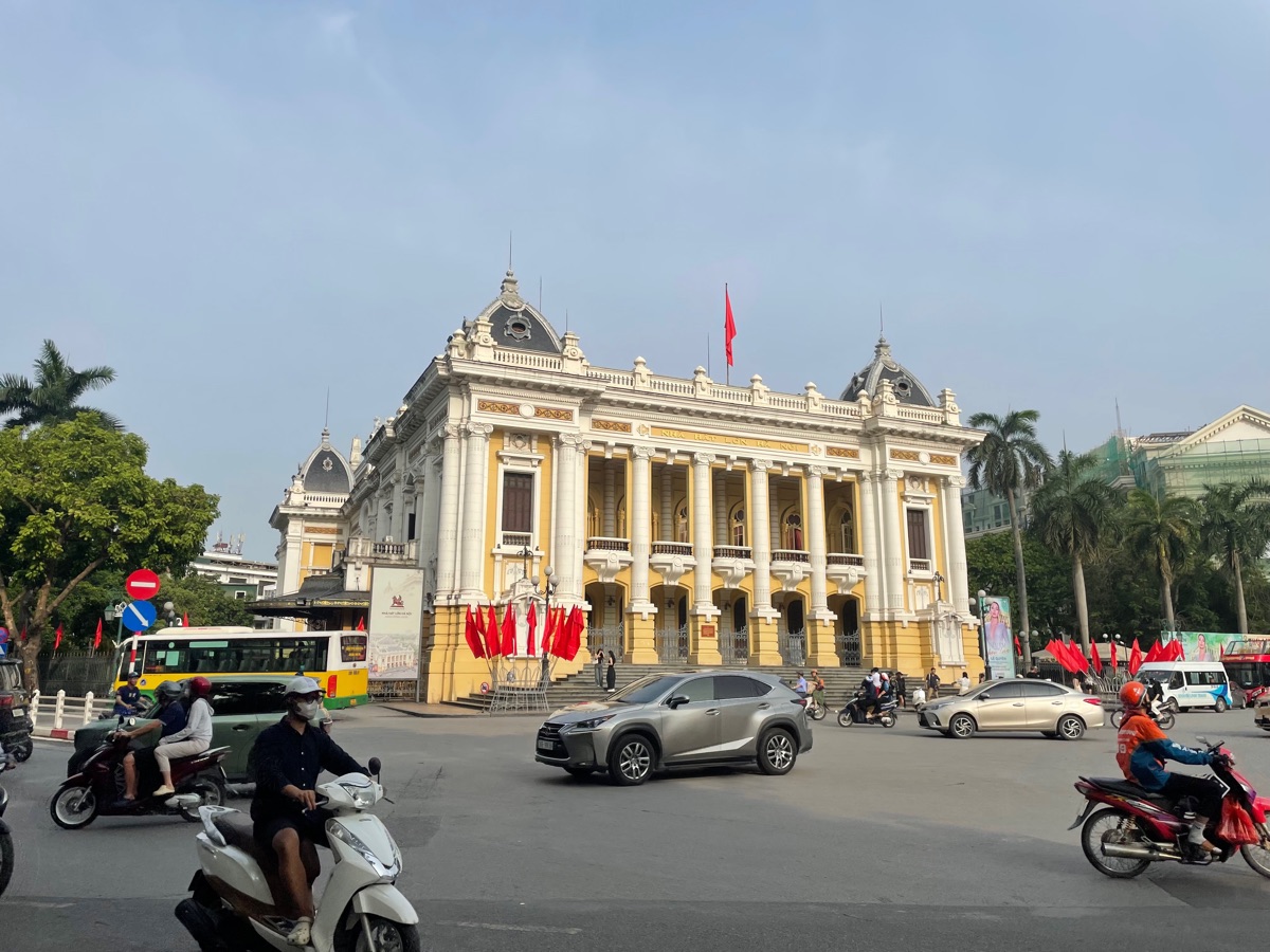 Het gele Operagebouw van Hanoi met rode vlaggen, en scooters op de weg ervoor