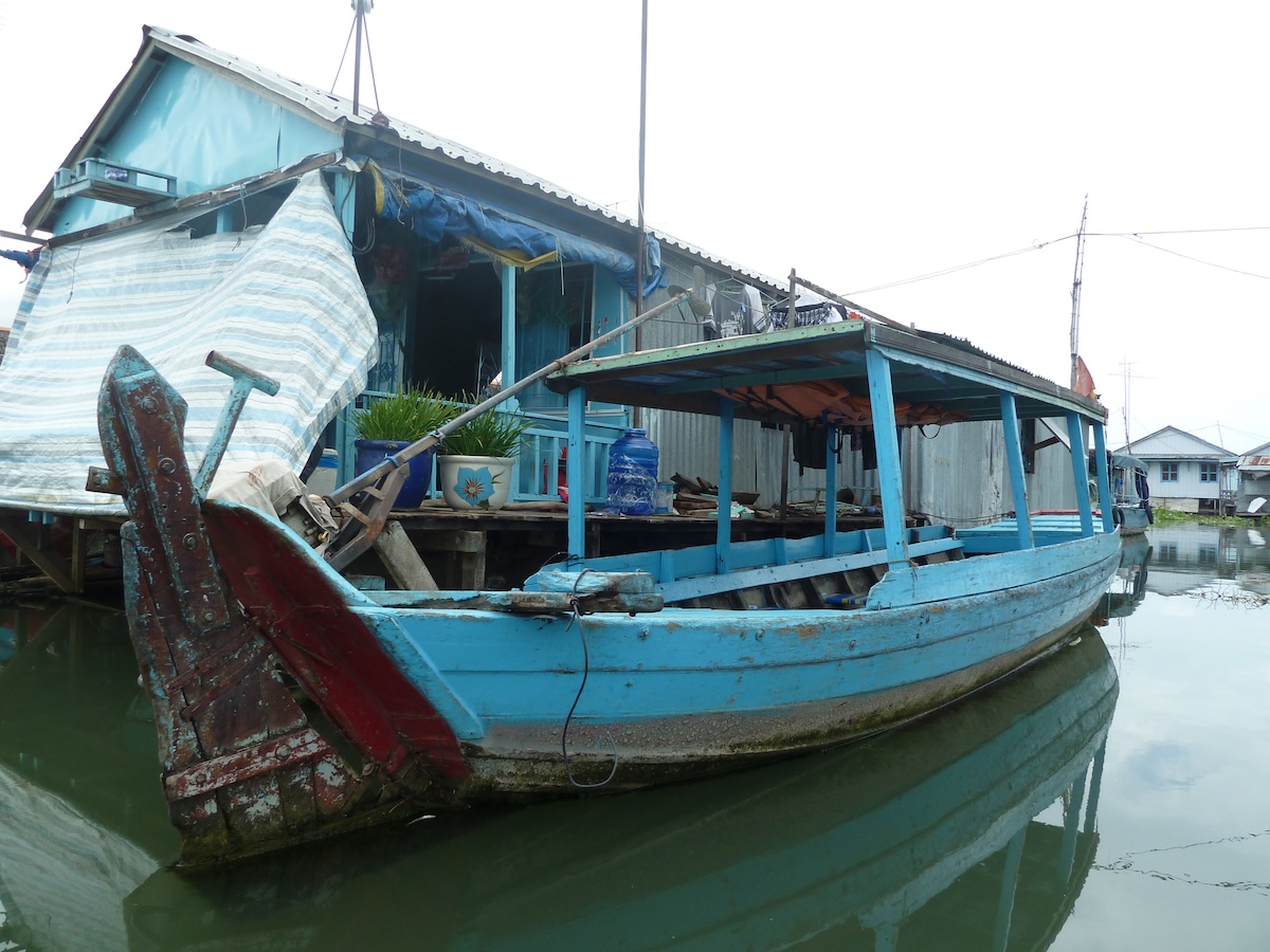 Een blauwe houten boot aangemeerd bij drijvende huizen op de rivier