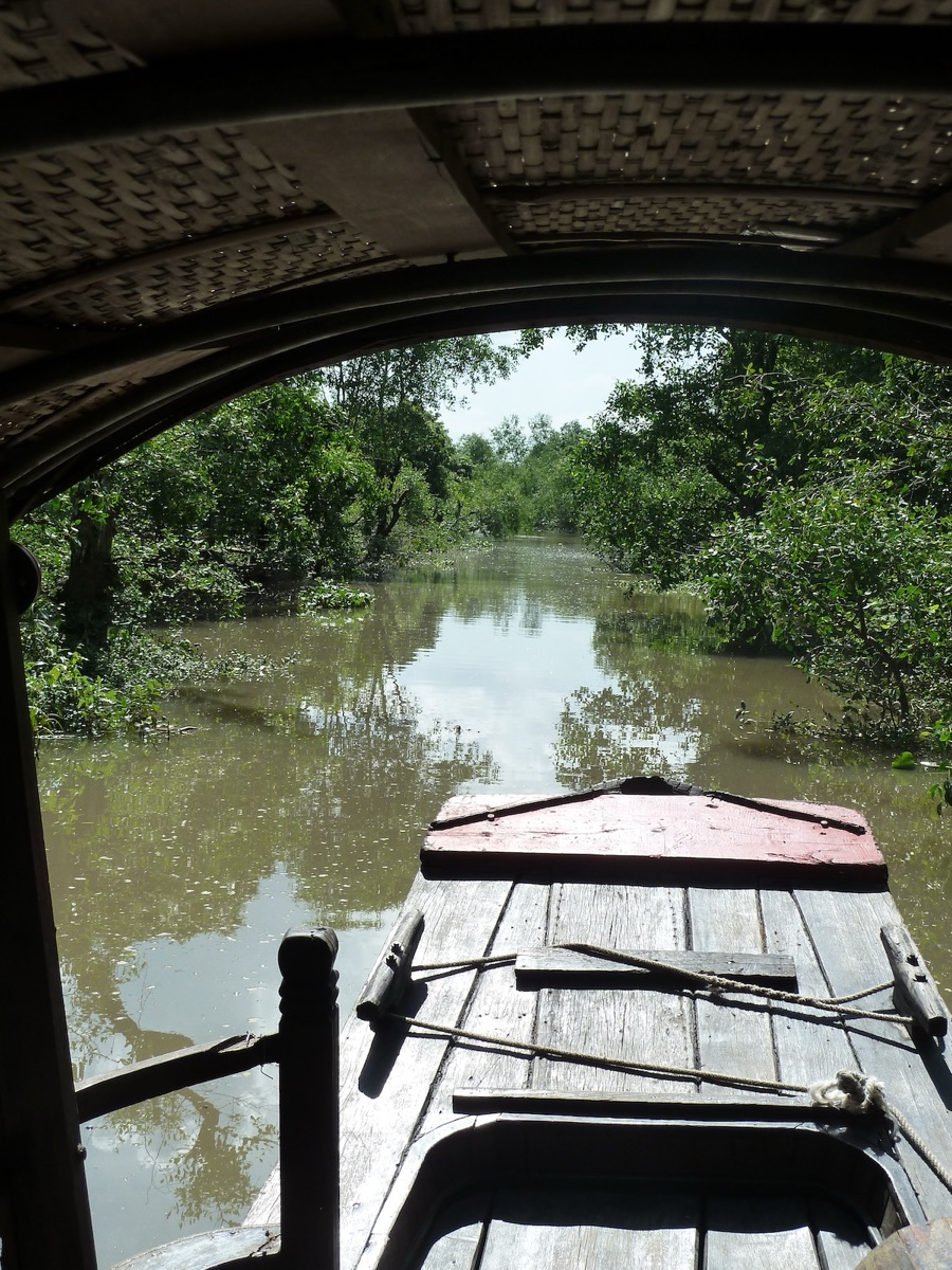 Uitzicht vanonder de overkapping van een houten boot over een smal kanaal tussen het groen