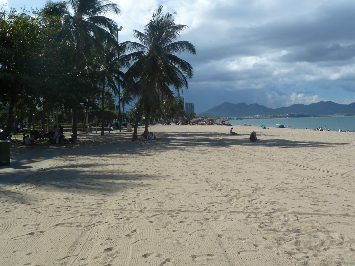 Het brede zandstrand van Nha Trang met palmbomen, en bergen aan de overkant van de baai